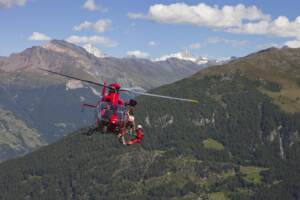 A MOUNTAIN RESCUE EMERGENCY PHYSICIAN IS HANGING BELOW A HELICOPTER HIGH ABOVE THE GROUND. A INJURED HIKER IS BEING WINCHED UP. THE SWISS ALPS IN THE BACKGROUND.