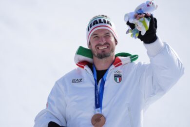 Dominik Paris festeggia il bronzo nella discesa libera maschile sollevando al cielo una delle due mascotte di queste Olimpiadi 2026