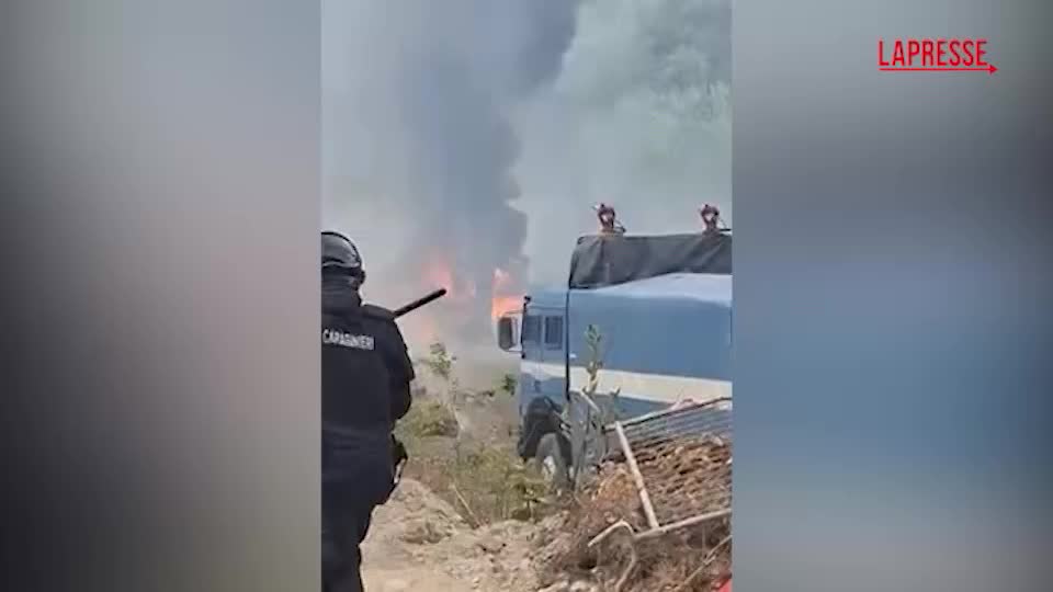 Fiamme all'interno di un cantiere durante manifestazione con manifestanti incappucciati e polizia in assetto di ordine pubblico